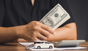 Man Holding Dollar Bill And Car Model On Desk, Representing Payday Loans, Title Loans, Loan Terms