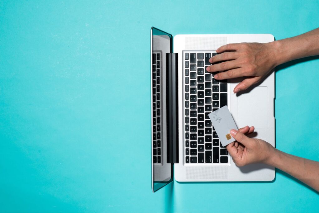 Credit card and laptop held in hands on turquoise surface, symbolizing finance