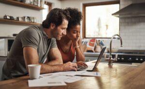 A man and woman sit at a kitchen table, looking at a laptop.