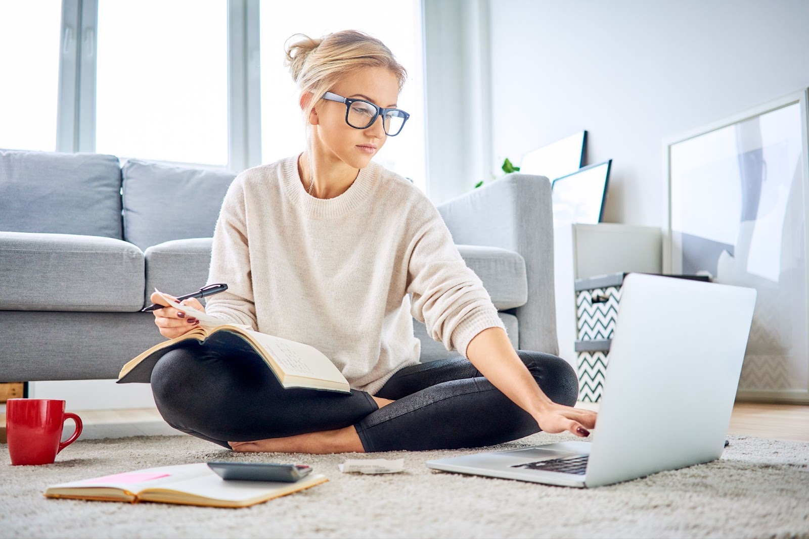 A woman in glasses sits on the floor, focused on her laptop in a cozy atmosphere.