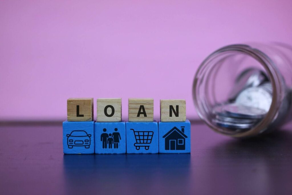 Four wooden blocks spelling LOAN sit atop four blue blocks featuring icons of a car, a family, a shopping cart, and a house, next to an overturned glass jar of coins.