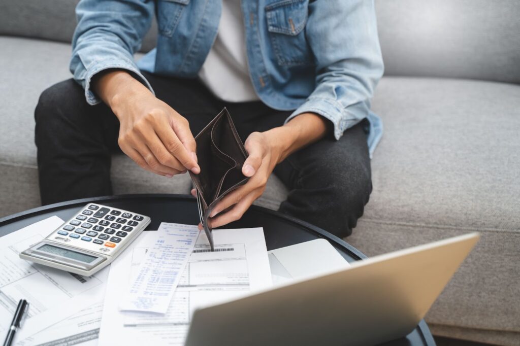 A man sitting on a couch, wearing a denim jacket, opening an empty wallet while looking at financial documents, a calculator, and a laptop on the table in front of him.