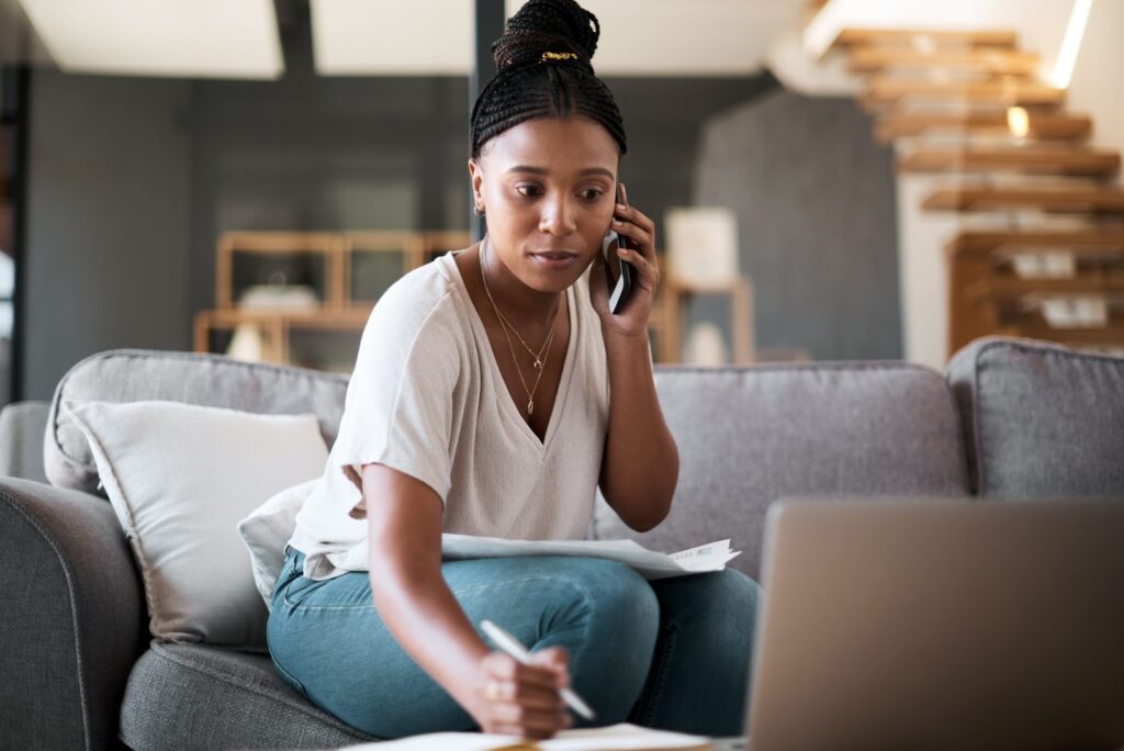 A woman sitting on a couch, talking on the phone while reviewing financial documents and taking notes on a piece of paper, with a laptop open in front of her.