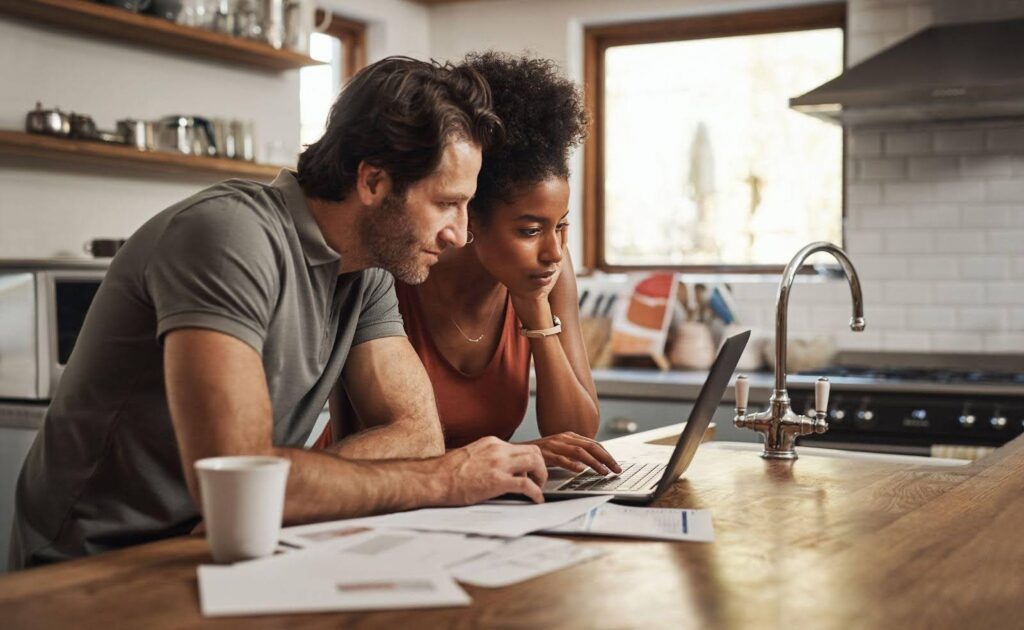 A couple at a kitchen table engaged in discussion while looking at a laptop together