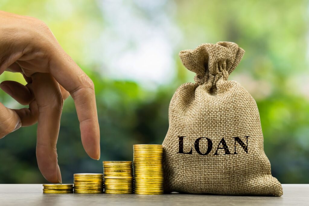 A hand stacks gold coins next to a burlap sack labeled LOAN on a table with a blurred green background.
