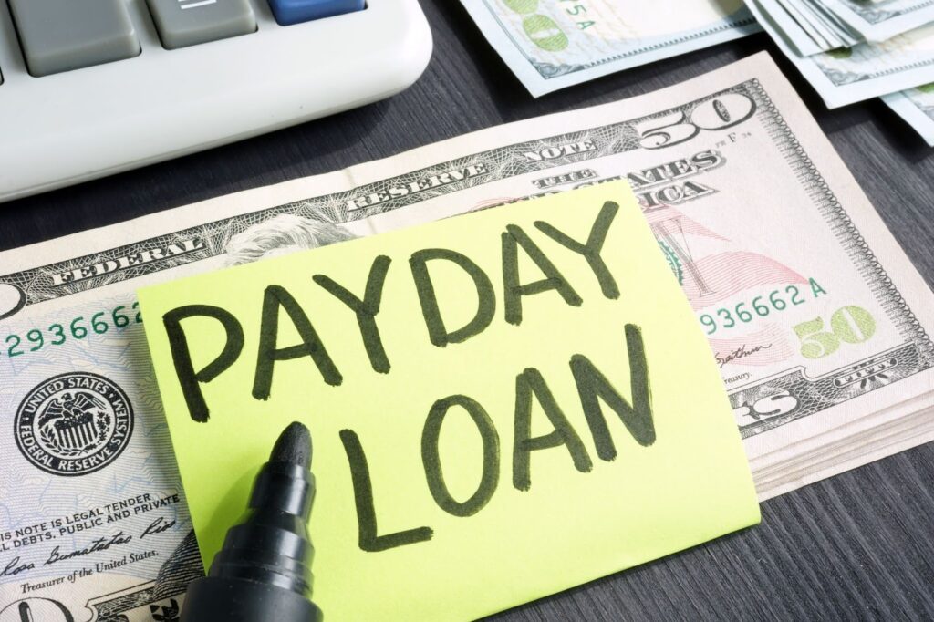 A yellow sticky note with PAYDAY LOAN written on it, placed on a stack of U.S. dollar bills next to a calculator.