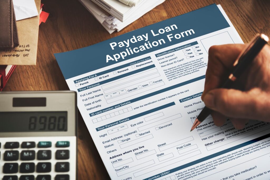 A hand fills out a Payday Loan Form on a desk with a calculator showing 8980, envelopes, and papers.