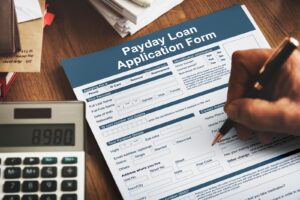 A hand fills out a Payday Loan Form on a desk with a calculator showing 8980, envelopes, and papers.
