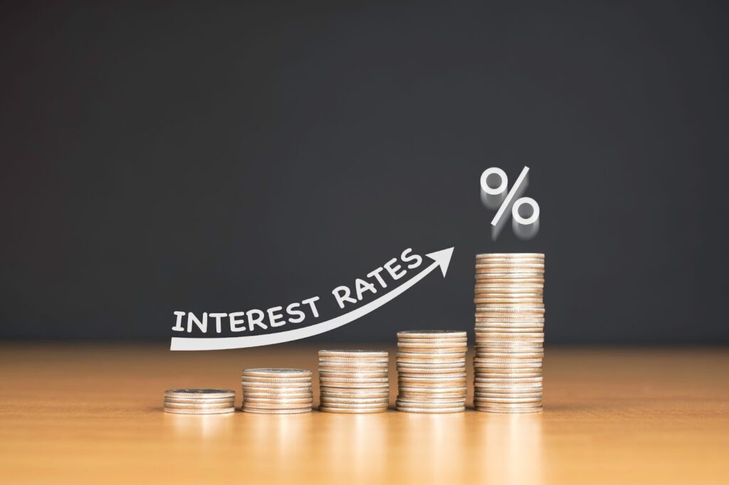 Stacks of coins in increasing height on a wooden surface with INTEREST RATES and a percentage symbol above, indicating rising rates.