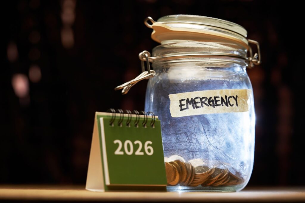 A glass jar labeled "EMERGENCY" containing coins sits next to a small green desk calendar displaying the year 2026.