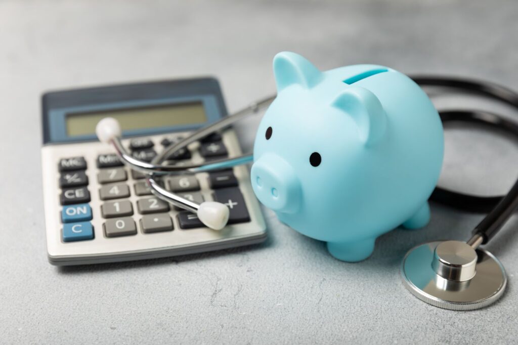 A light blue piggy bank, a stethoscope, and a calculator on a gray surface, symbolizing healthcare costs or medical savings.