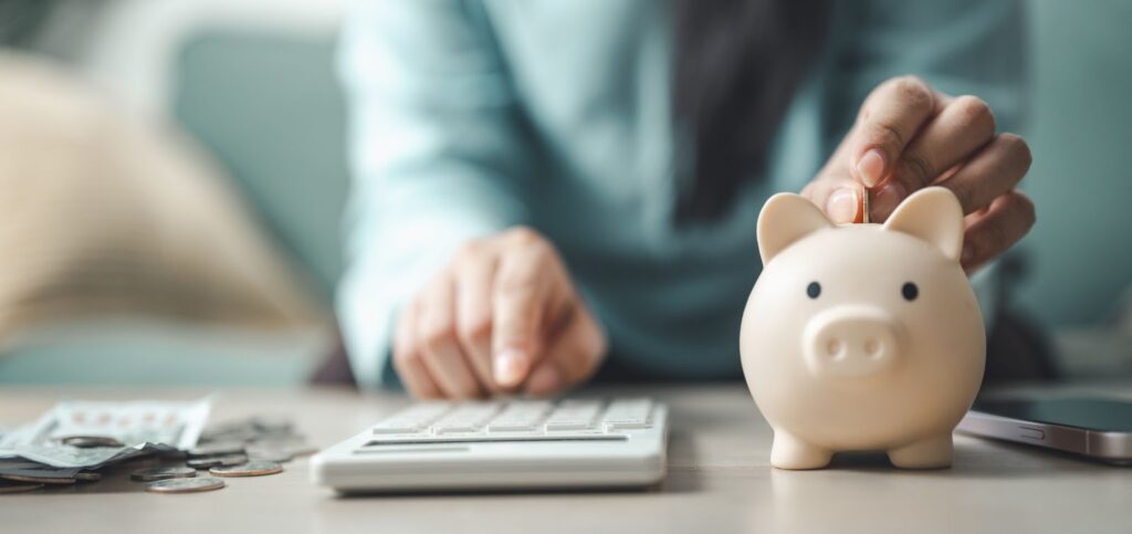 A person placing a coin into a beige piggy bank while using a calculator, with cash and coins nearby on a desk.