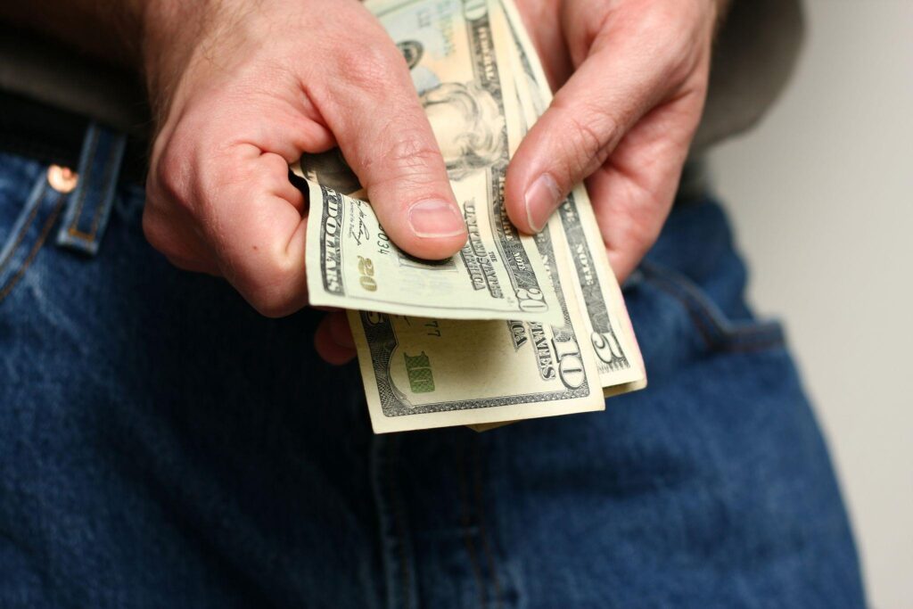 A person in jeans holds a small stack of U.S dollar bills displaying denominations of five and ten