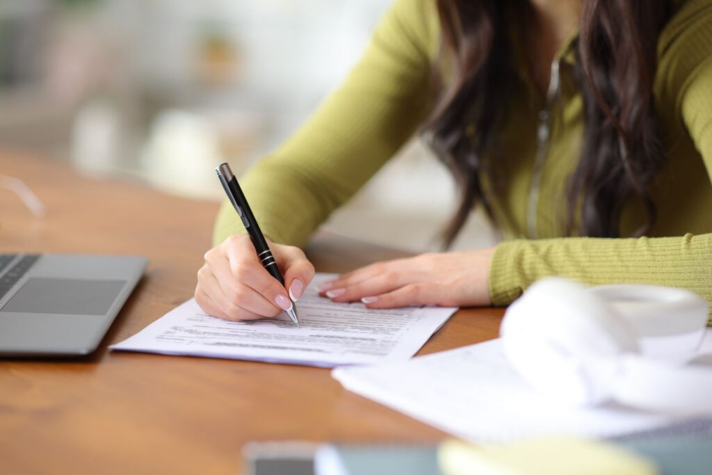 Close-up of a woman's hands filling out a printed form with a black pen at a wooden desk next to a laptop.