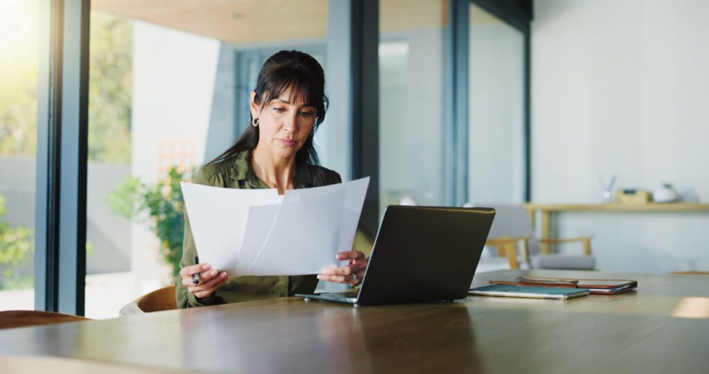 Professional woman reading printed documents while sitting at a table with an open laptop in a bright home office.